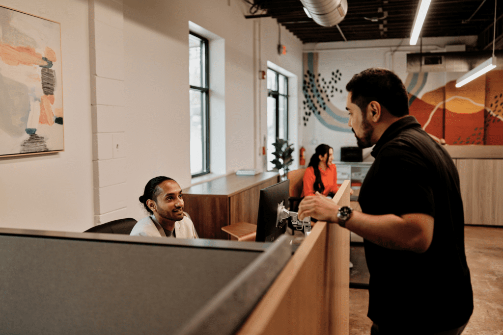 Two colleagues chat around a cubicle in an office.